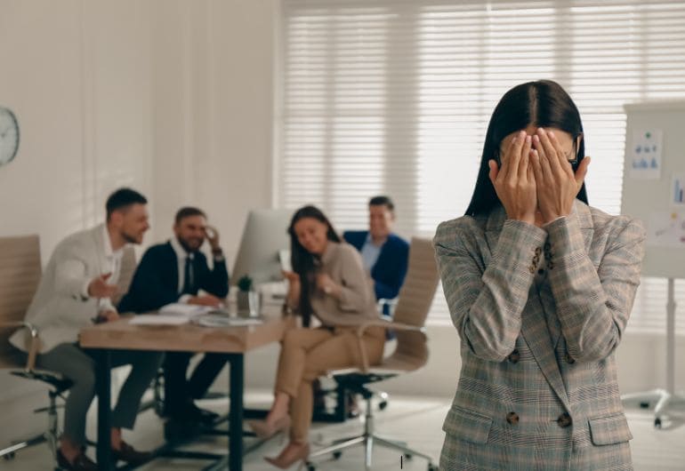 Woman feeling excluded in office while others laugh, symbolizing unfair treatment. Text: "How ‘No Unfair Helping’ Rules Benefit Small Shops | Tech Garcons."